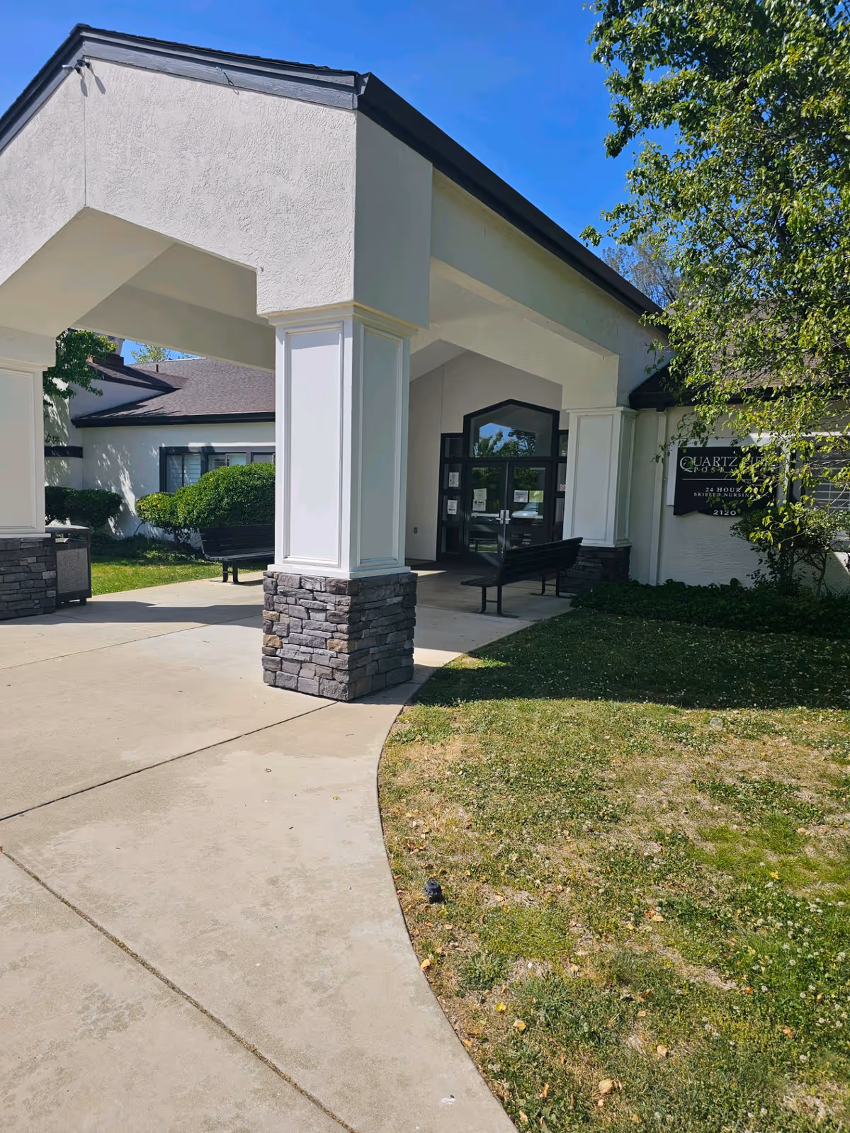 Entrance to Quartz Hill Post Acute facility with a covered walkway supported by white columns with stone bases, benches near the entrance, green bushes, and a tree on the right side under a clear blue sky.