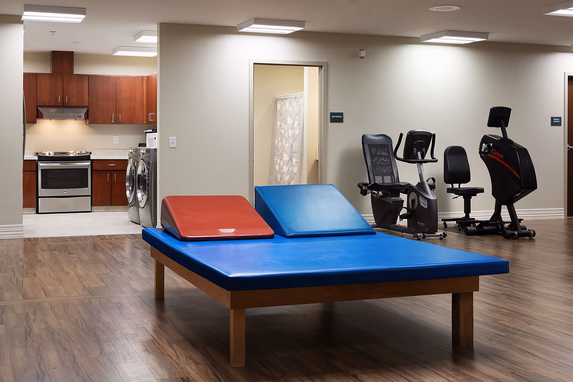 Interior view of a therapy or exercise room with a large blue therapy table with red and blue angled cushions in the center. To the right, there are three exercise machines including a recumbent bike and a seated elliptical. In the background, there is a doorway leading to a bathroom with a shower curtain visible. To the left, a kitchen area with wooden cabinets, a stove, and a washer and dryer is visible. The room has wood flooring and neutral-colored walls.