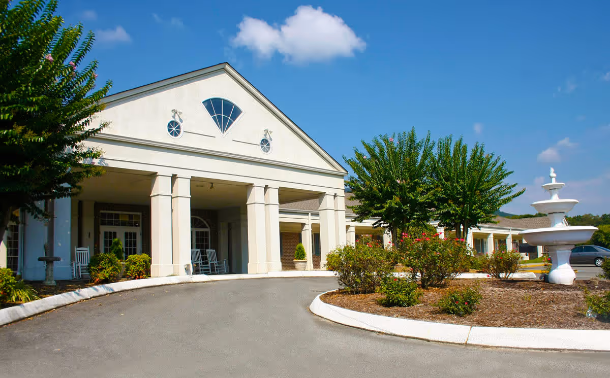 Front exterior view of a senior living facility with a white building featuring large columns, a covered entrance with rocking chairs, landscaped bushes and trees, and a white tiered fountain on the right side under a blue sky with some clouds.