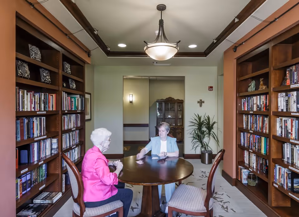 Two elderly women sitting at a round wooden table playing cards in a cozy library room with bookshelves filled with books on both sides. The room has a ceiling light fixture, a potted plant, and a wooden cabinet in the background.