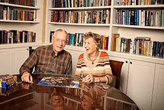 An elderly man and woman sitting at a glass table in a room with white built-in bookshelves filled with books. They are smiling and appear to be working on a jigsaw puzzle together.