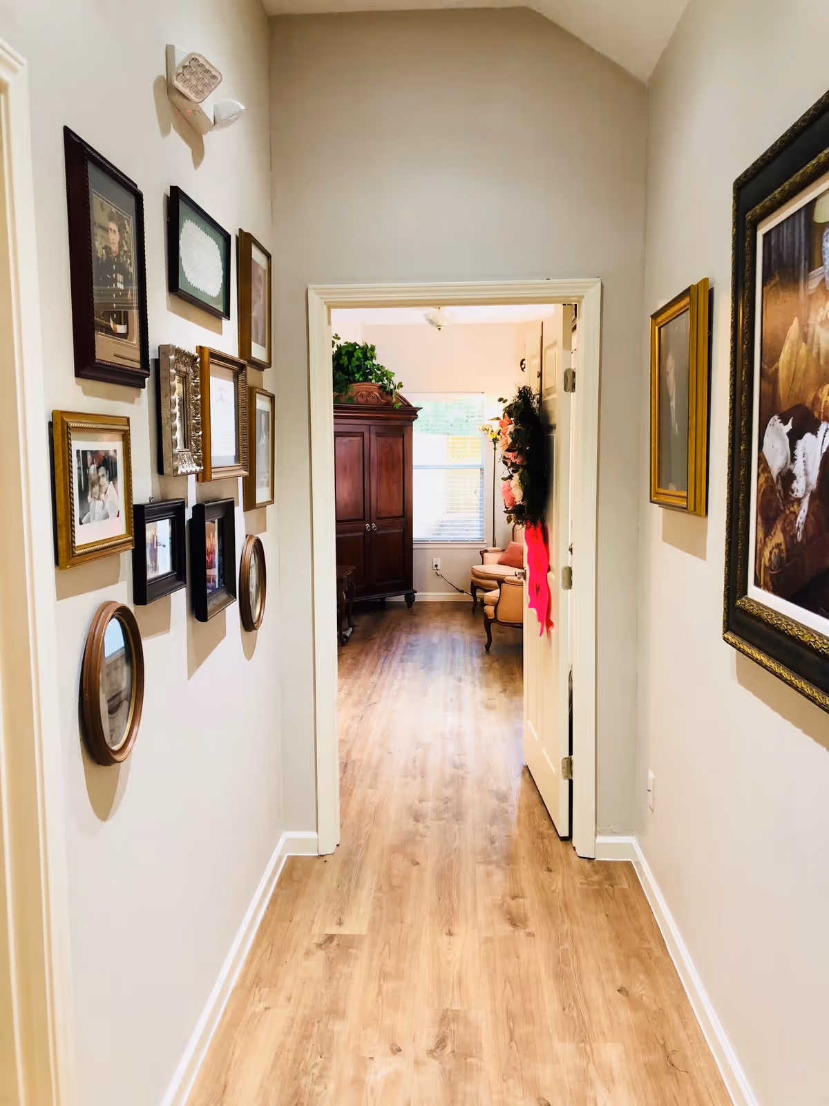 A hallway with light-colored walls and wooden flooring leading to a room with a large wooden cabinet, a window with blinds, and a couple of upholstered chairs. The hallway walls are decorated with various framed pictures and artwork.