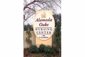 A tall beige sign with a rounded top displaying the name 'Alameda Oaks Nursing Center' along with the address '1100 S. Alameda'. The sign is set outdoors with some greenery and a tree in the background.
