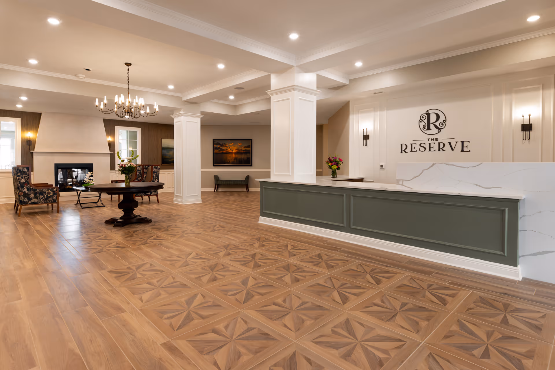 Spacious and elegant lobby area of The Reserve at Fairhope featuring a patterned wooden floor, a green and white reception desk with a marble countertop, a round wooden table with a flower vase, two floral upholstered chairs near a fireplace, a chandelier, and wall sconces. The wall behind the reception desk displays the logo and name 'The Reserve'.