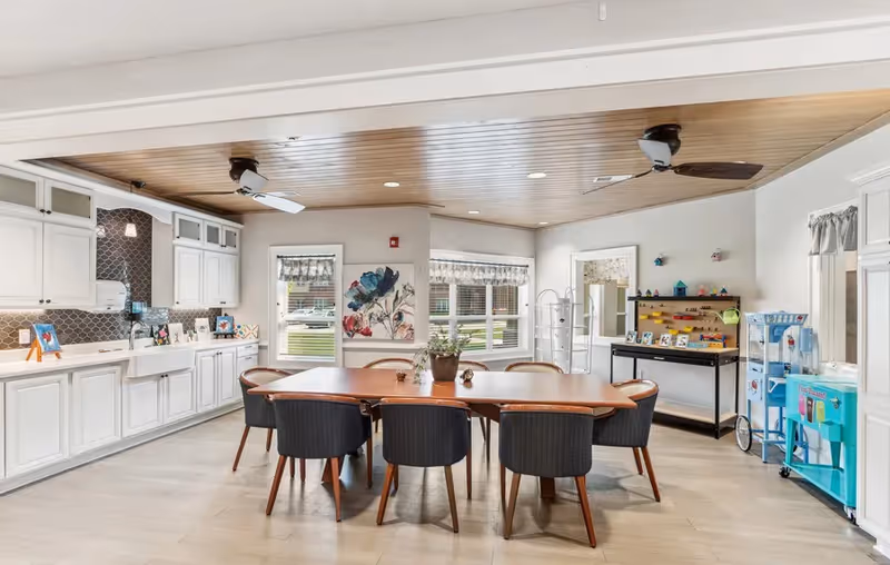 A bright and spacious kitchen and dining area with white cabinetry, a farmhouse sink, and a wooden dining table surrounded by eight dark cushioned chairs. The ceiling has wood paneling with two ceiling fans. Large windows with valances let in natural light. There are decorative items on the countertops and a small display shelf with various items on the right side of the room.