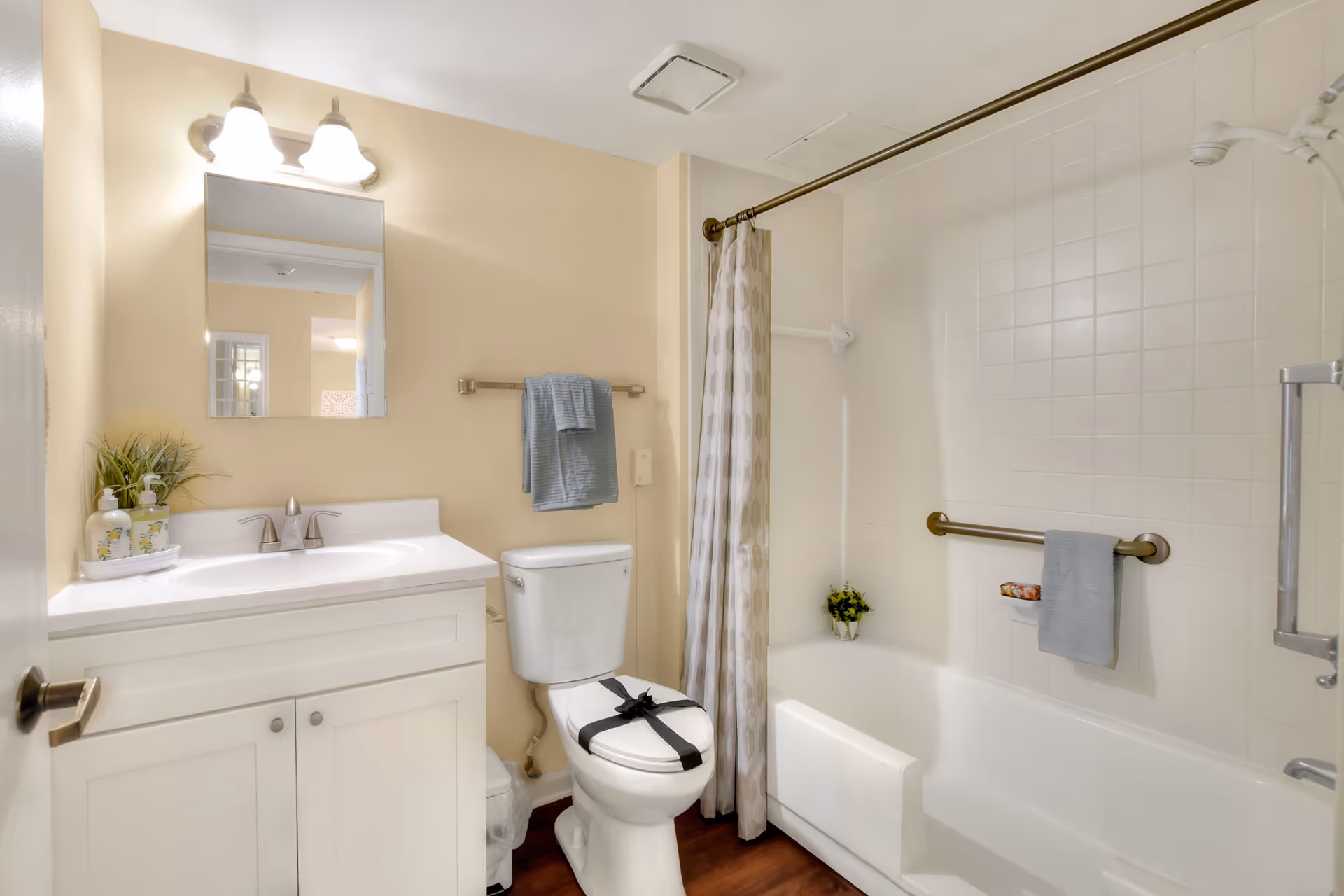 Bright bathroom with a white vanity and mirror, toilet, and a tub-shower with tiled surround, curtain, and grab bars.