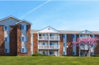 Front view of a three-story brick building with balconies, a green lawn, and a flowering tree under a blue sky.