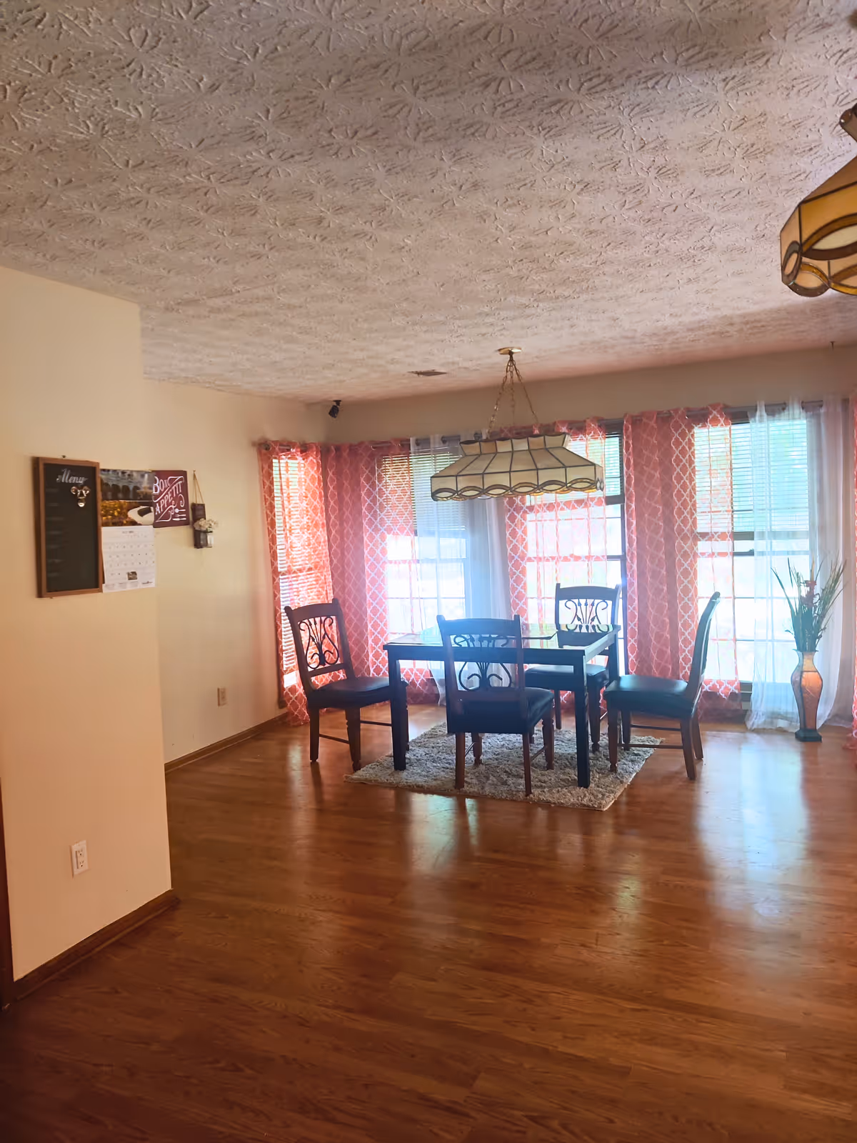 Dining room with a wooden table and chairs under a hanging light in front of large windows with red patterned curtains.