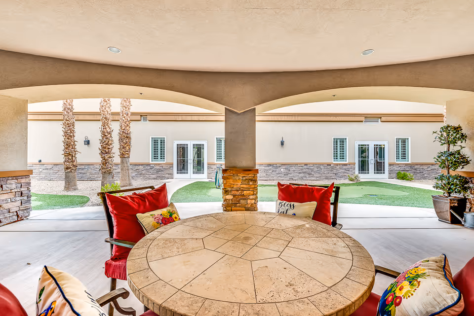 Covered outdoor patio area with a round stone table surrounded by chairs with red cushions and decorative pillows. The patio overlooks a courtyard with green artificial grass, palm trees, potted plants, and a beige building with windows and doors in the background.