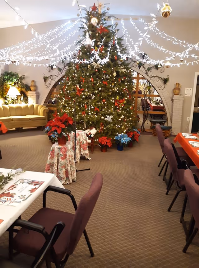 Decorated common room with a large Christmas tree, string lights overhead, and tables and chairs set for dining.