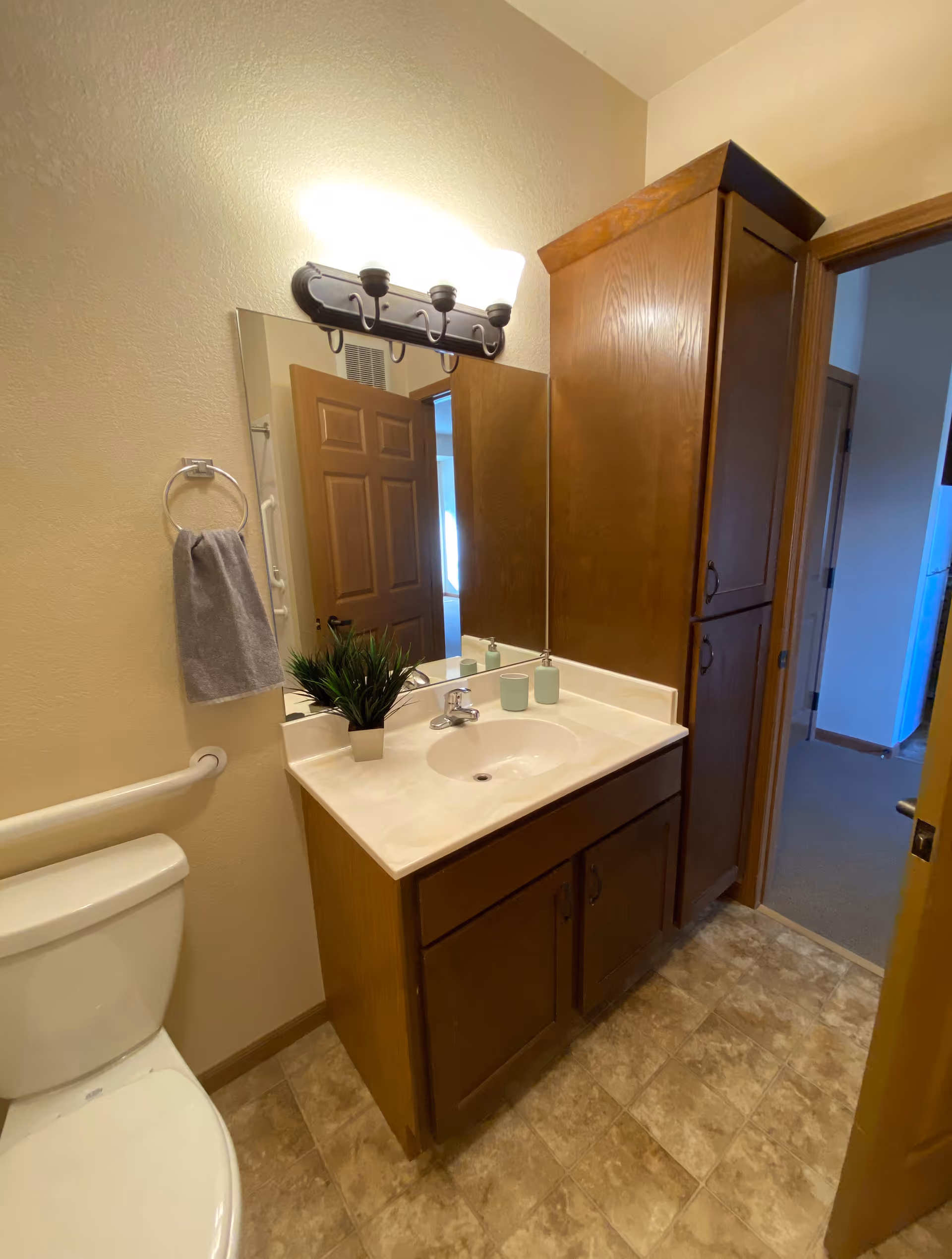 A bathroom with a white toilet, a wooden vanity with a white countertop and sink, a large mirror above the sink, a wall-mounted light fixture with three bulbs, a gray hand towel hanging on a ring, and a tall wooden cabinet next to the vanity. The floor has beige and brown tiles, and an open door leads to a carpeted room.
