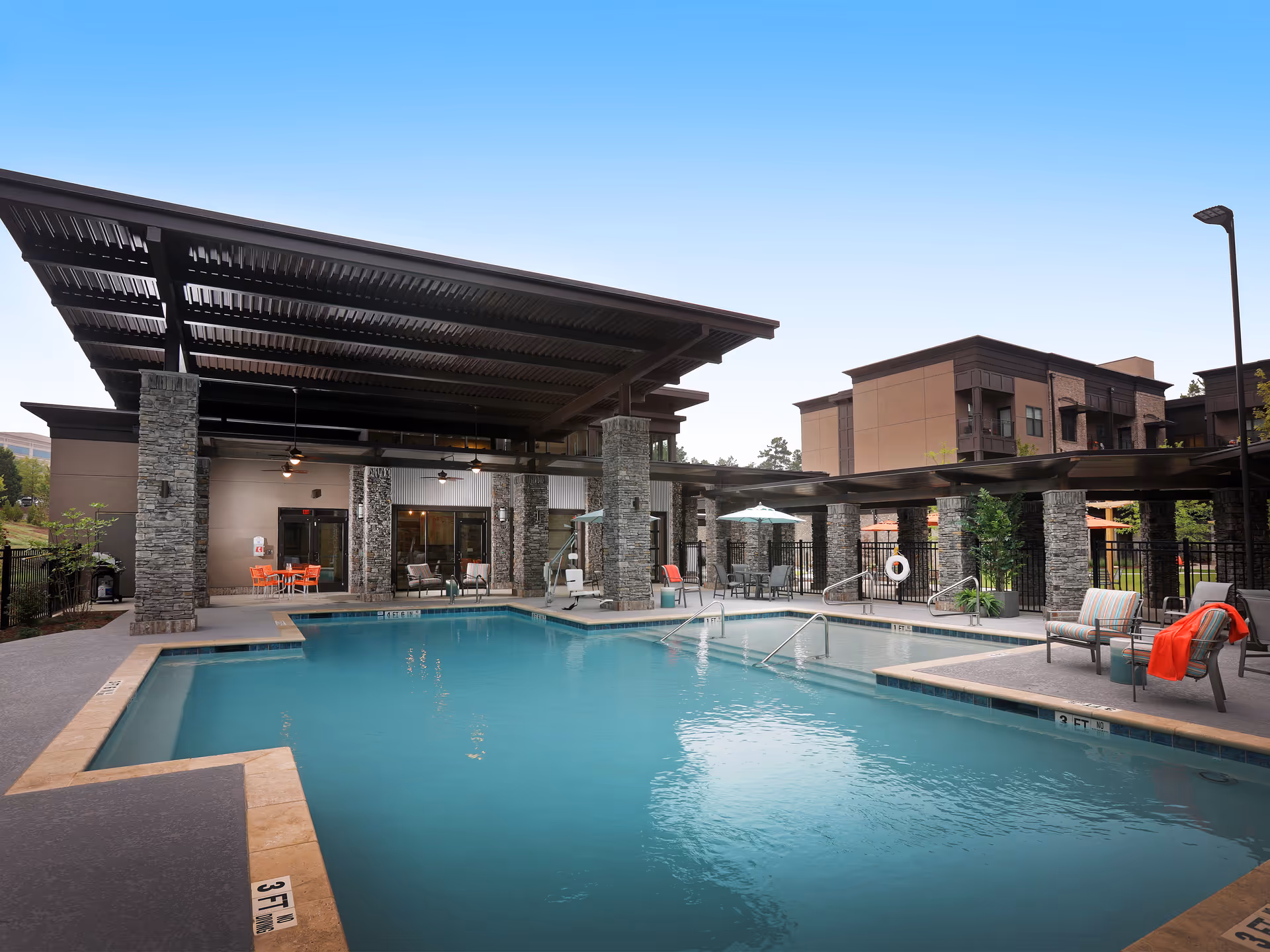 Outdoor swimming pool area at Village Park Alpharetta with a covered seating area supported by stone pillars, lounge chairs with cushions and a red blanket, tables with umbrellas, and a clear blue sky.