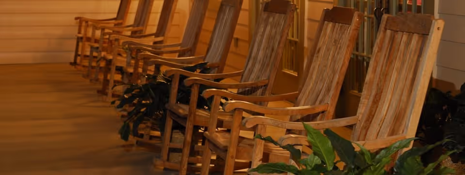 A row of wooden rocking chairs lined up on a covered porch with potted plants and warm lighting.
