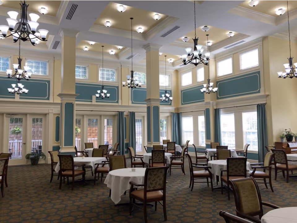 A spacious dining room with multiple round tables covered with white tablecloths and surrounded by wooden chairs with cushioned seats and backs. The room features tall ceilings with elegant chandeliers and recessed lighting. Large windows and glass doors allow natural light to fill the space. The walls are painted in a soft yellow with teal panel accents, and there are decorative columns throughout the room.