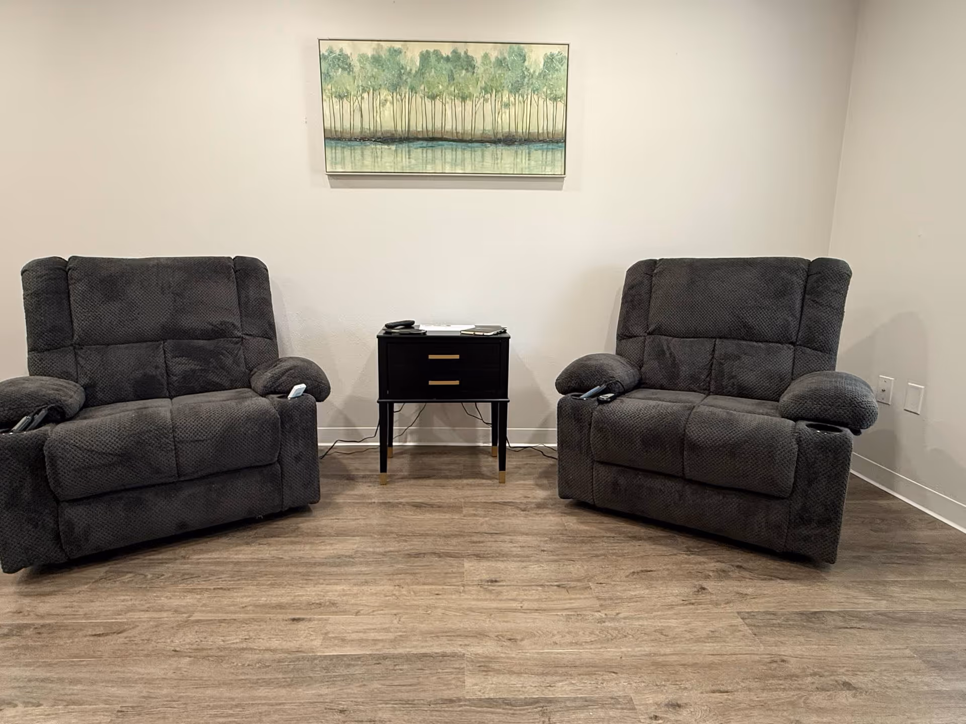 Two dark gray recliner chairs with armrests and cup holders are positioned on either side of a small black side table with two drawers. The table has some items on top, including a remote control. Above the table, a framed painting of a forest with green trees and a reflective water body hangs on a plain white wall. The floor is wooden with a light brown finish.