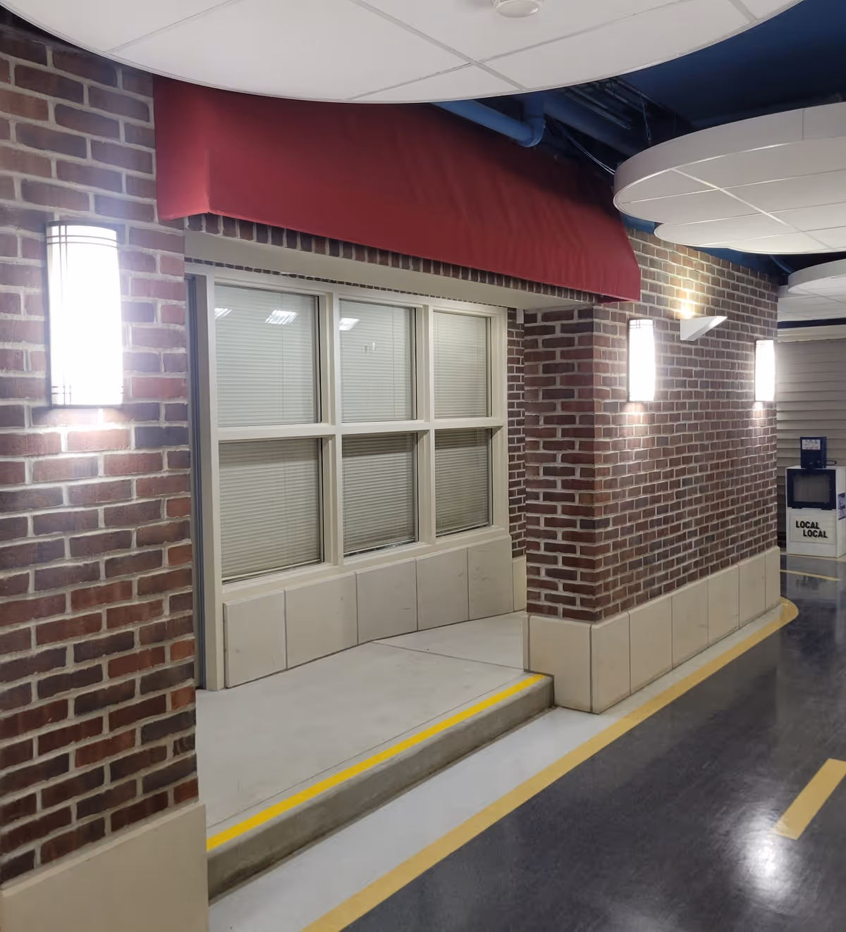 Indoor hallway with brick walls, a window with closed blinds, a red fabric awning above the window, and three wall-mounted lights illuminating the area. The floor has a yellow line marking a step down from the window area to the hallway. Ceiling panels and exposed pipes are visible above.