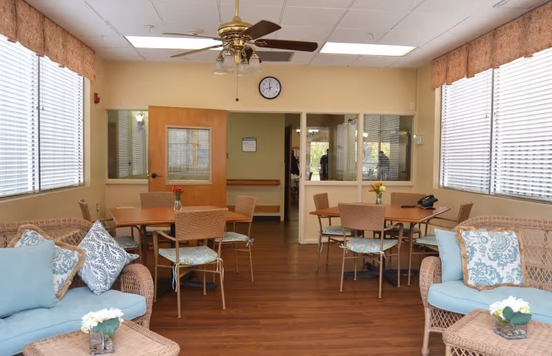 Bright communal lounge and dining area with wicker sofas, multiple tables and chairs, large windows and a ceiling fan.