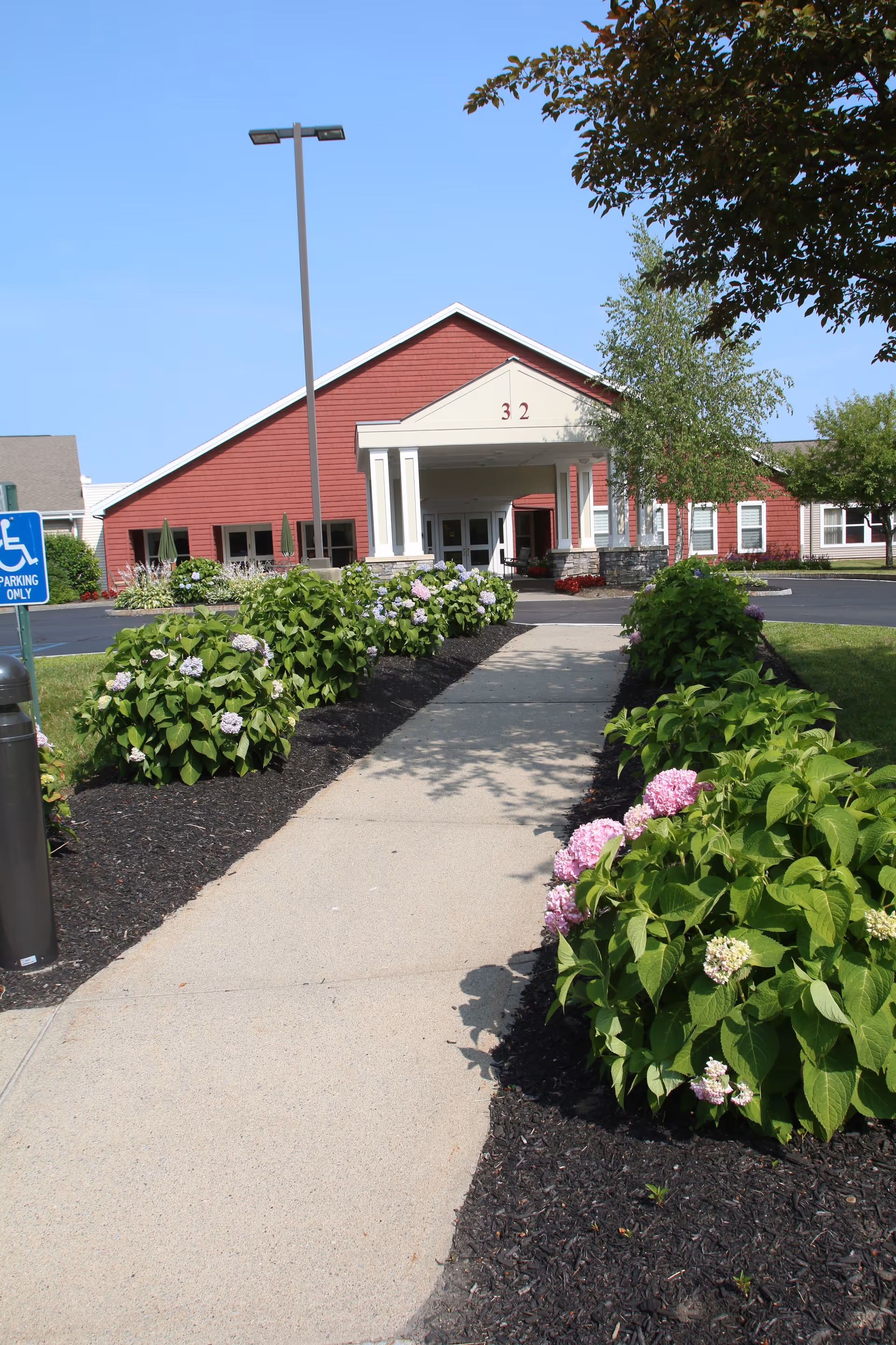 A concrete walkway lined with green bushes and blooming flowers leads to the entrance of a red building with white trim and the number 32 above the entrance. There is a tall street lamp on the left side and a handicapped parking sign visible near the walkway. The sky is clear and blue.