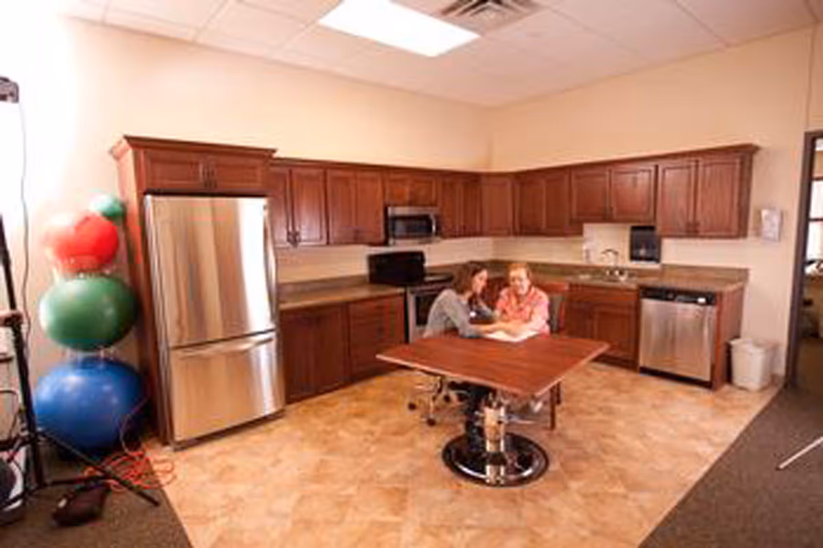 A kitchen area with wooden cabinets, a stainless steel refrigerator, microwave, stove, and dishwasher. Two women are sitting at a table in the center of the room, engaged in conversation or an activity. There are colorful exercise balls stacked in the corner to the left.