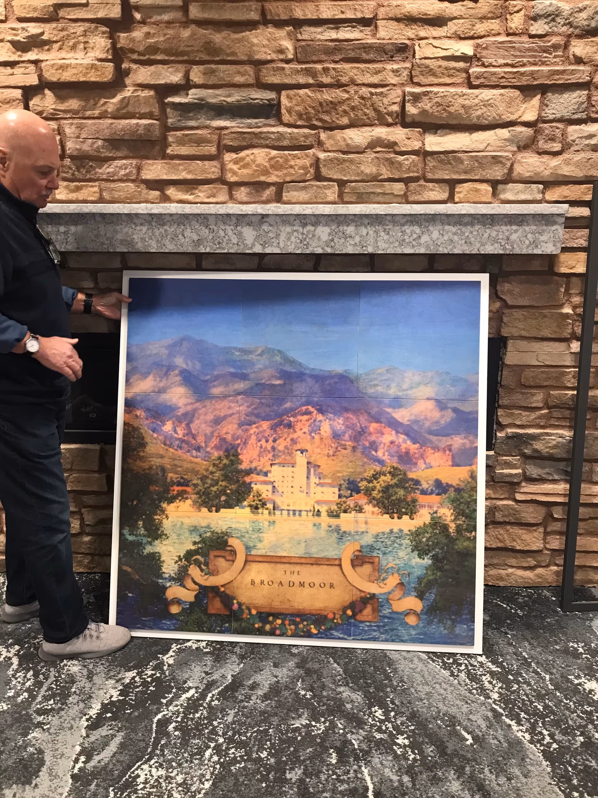An elderly man standing indoors next to a large framed picture depicting a scenic view of mountains, a lake, and a building labeled 'The Broadmoor'. The background features a stone wall and a gray stone mantel above a fireplace.
