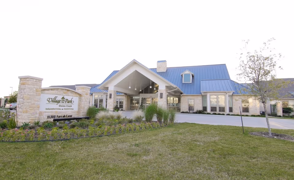 Exterior front view of Village on the Park Onion Creek senior living facility with a stone sign displaying the facility name and address, a covered entrance, and landscaped lawn with plants and trees.