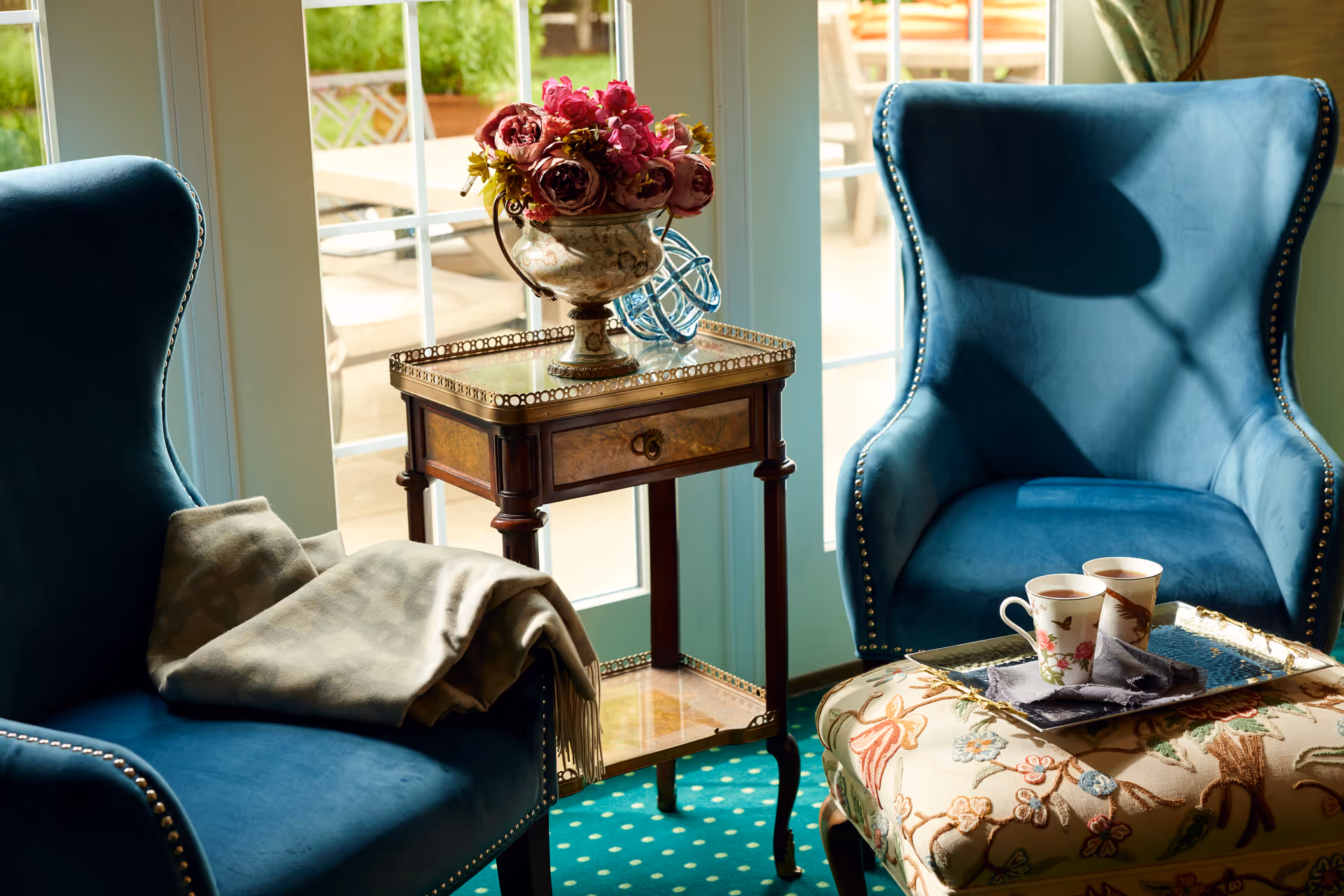 Two blue upholstered armchairs with nailhead trim positioned near a glass door leading outside. Between the chairs is a small wooden side table with a decorative vase of pink flowers and a blue glass ornament. One chair has a folded beige blanket draped over the armrest. In front of the other chair is a floral-patterned ottoman with a tray holding two floral teacups and a napkin.