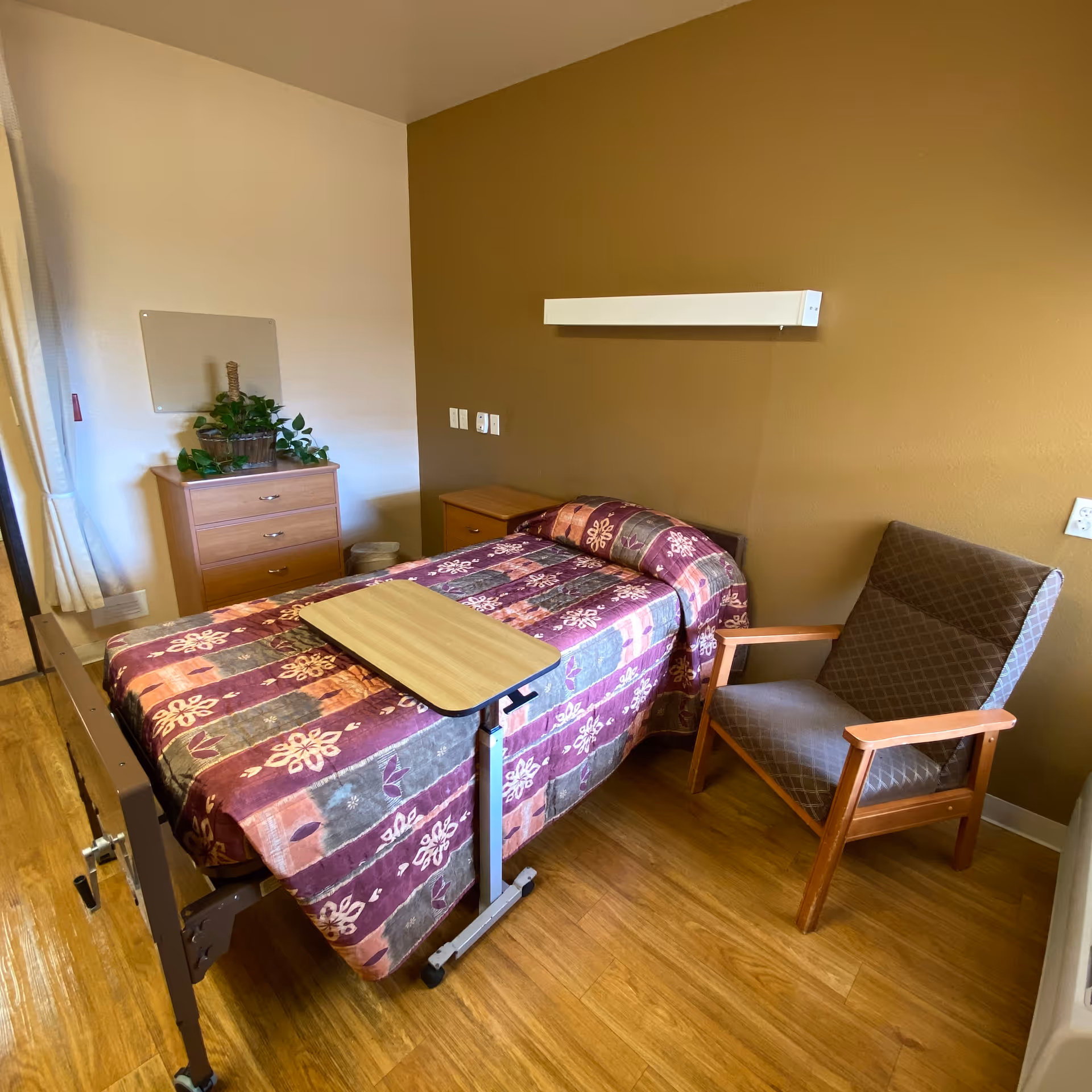 A small nursing facility bedroom with a single bed covered in a patterned quilt, a wooden bedside table, a wooden chest of drawers with a potted plant on top, and a cushioned armchair with wooden arms. The walls are painted in two tones, white and brown, and the floor is wooden.