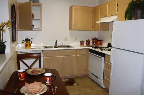 Small furnished kitchen with light wood cabinets, white refrigerator and stove, a sink, and a table set for two.