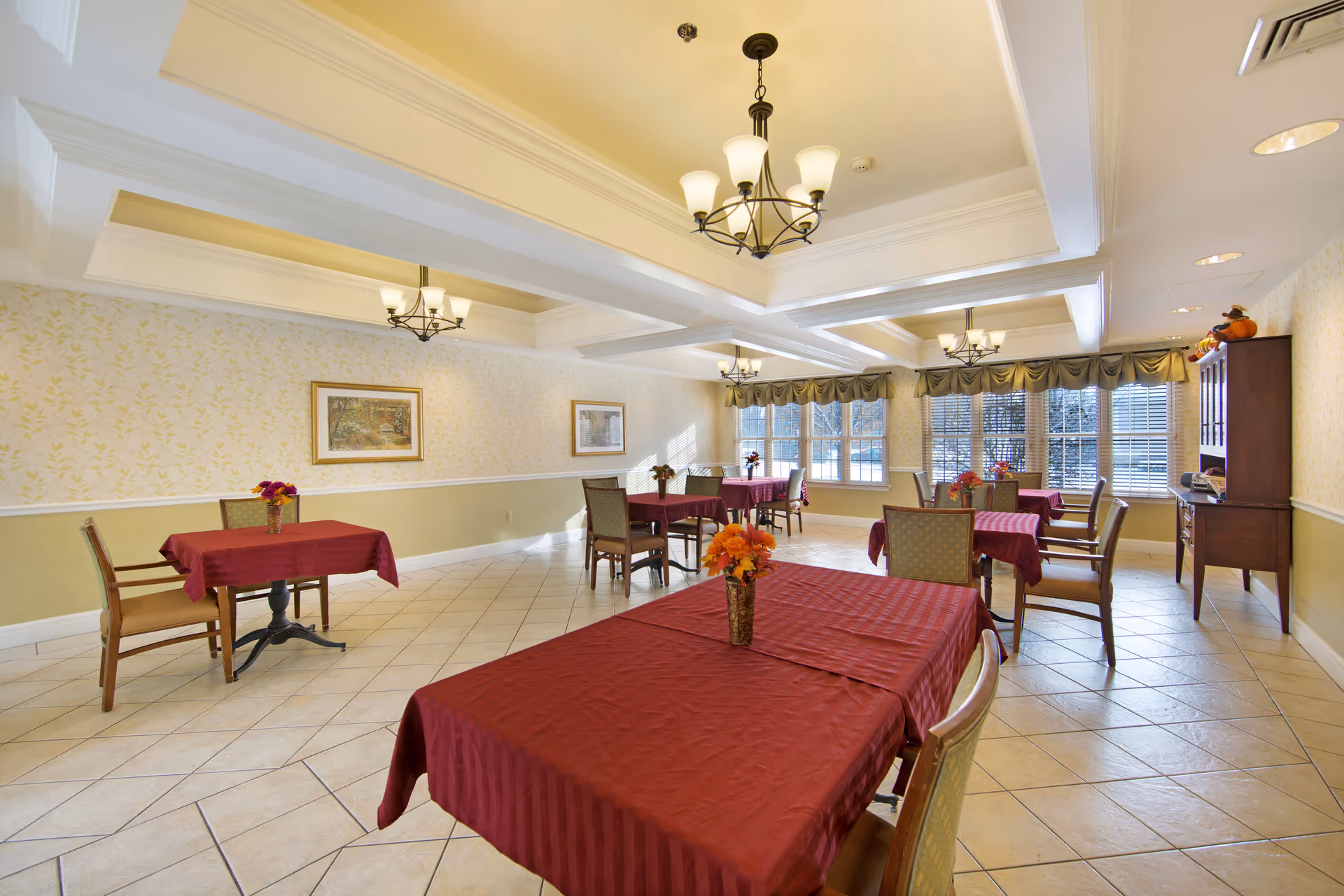 A bright dining room with several tables covered in red tablecloths, each adorned with a small vase of flowers. The room has large windows with blinds and valances, light-colored tiled floors, and multiple chandeliers hanging from a coffered ceiling. There are framed pictures on the walls and a wooden cabinet in the corner.