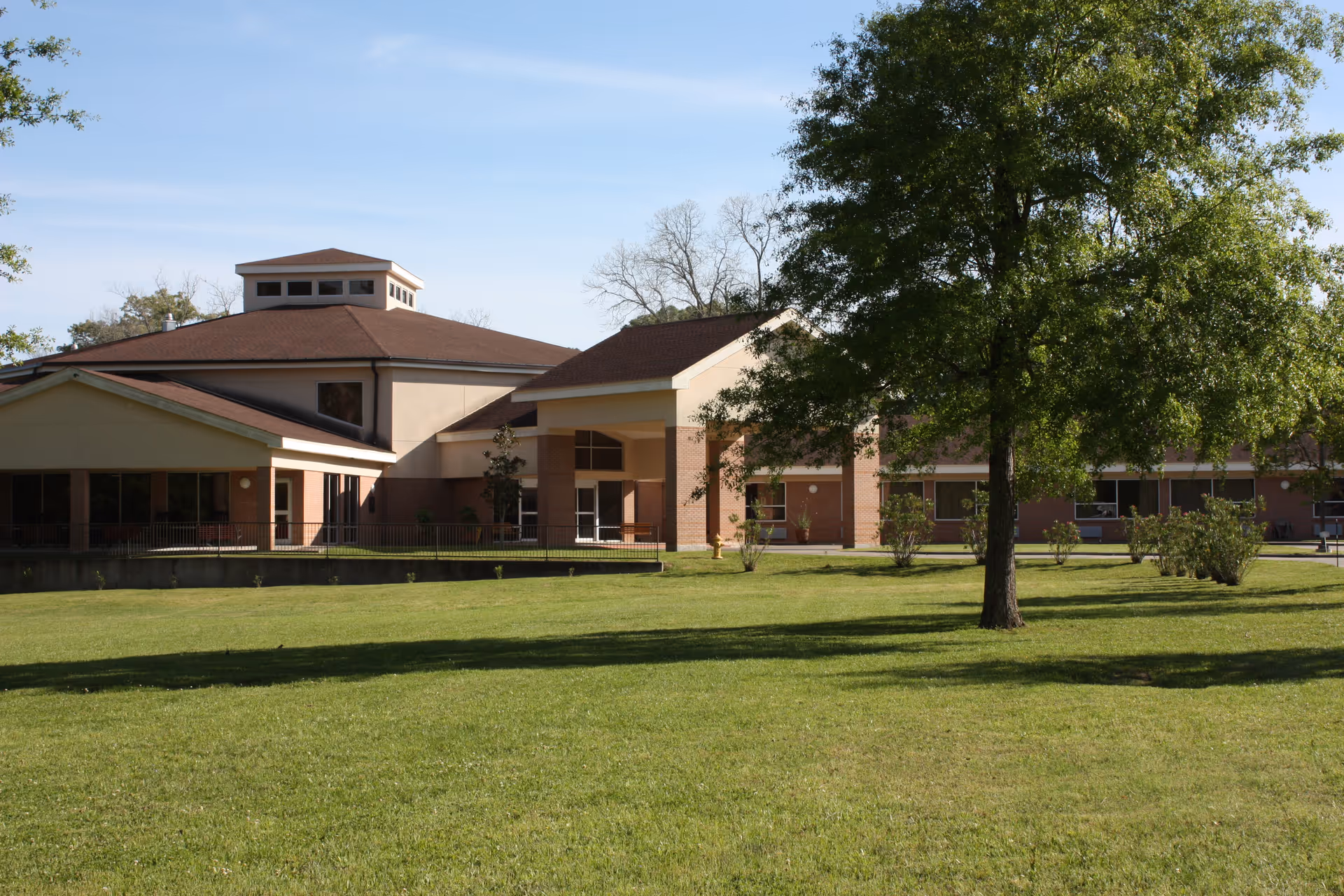 Exterior view of a single-story nursing facility and independent living center building with a brown roof and beige walls, surrounded by a well-maintained green lawn and trees under a clear blue sky.
