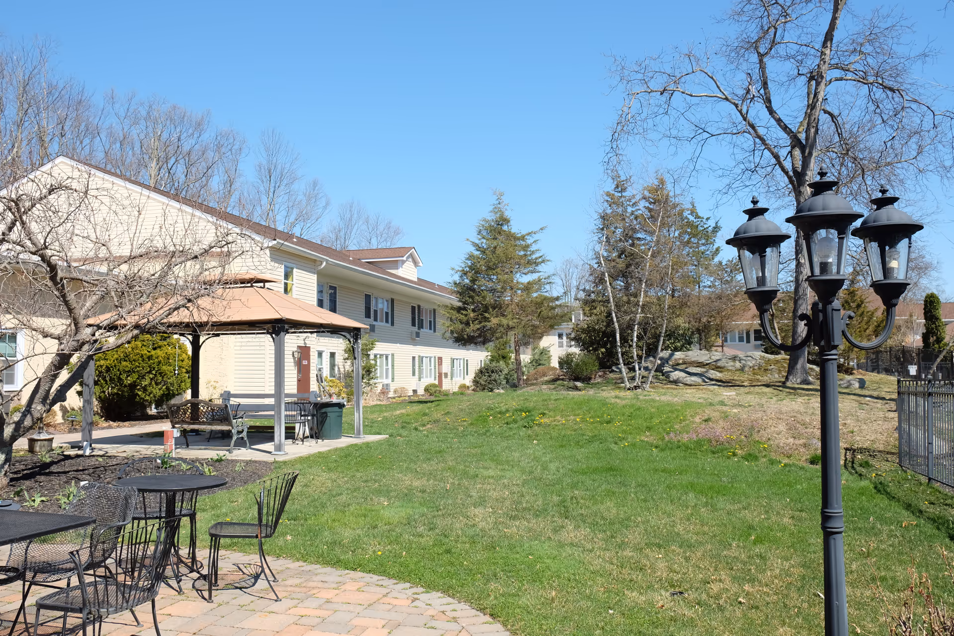 Outdoor garden area of a senior living facility with a paved patio featuring black metal tables and chairs, a gazebo with benches, a grassy lawn, trees, and a black vintage-style street lamp under a clear blue sky.