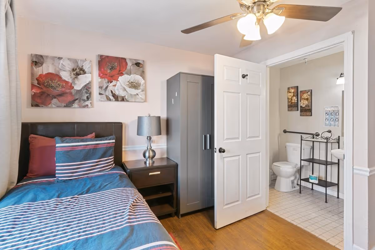 A bedroom with a single bed featuring striped bedding and pillows, a nightstand with a lamp, a gray wardrobe, and an open door leading to a bathroom with a toilet, sink, and decorative wall art.