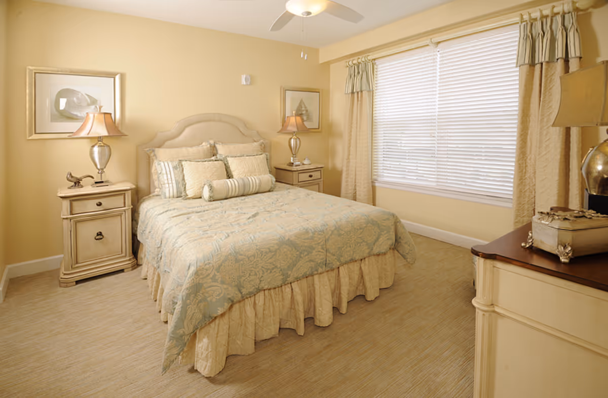 A cozy bedroom with a neatly made bed featuring a patterned bedspread and multiple pillows. The room has beige walls, carpeted floor, two matching bedside tables with lamps, framed artwork on the walls, a ceiling fan, and a large window with blinds and curtains.