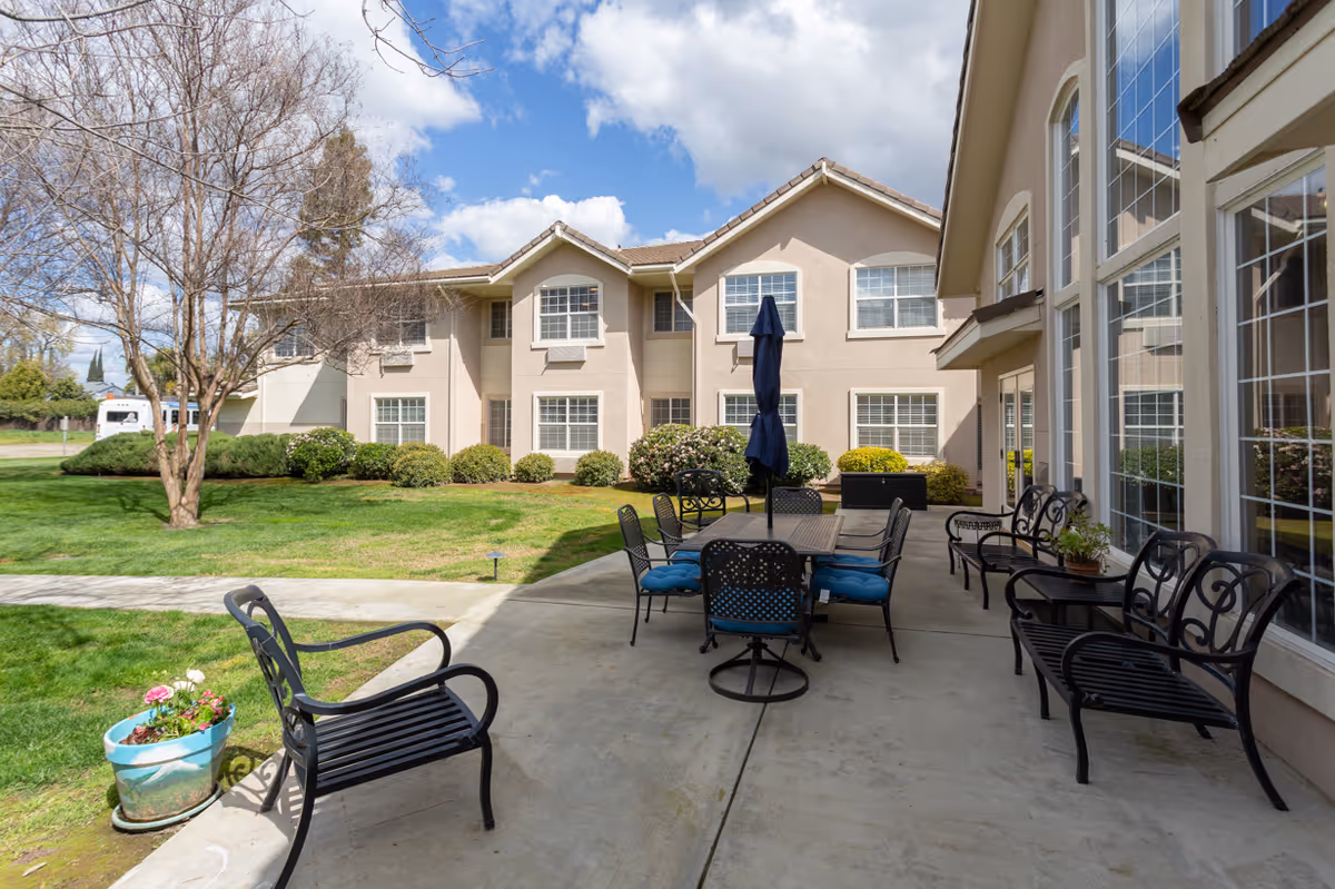 Outdoor patio area at Marbella Visalia with metal chairs and a table with a closed umbrella, surrounded by a well-maintained lawn, bushes, and a two-story beige building under a partly cloudy sky.