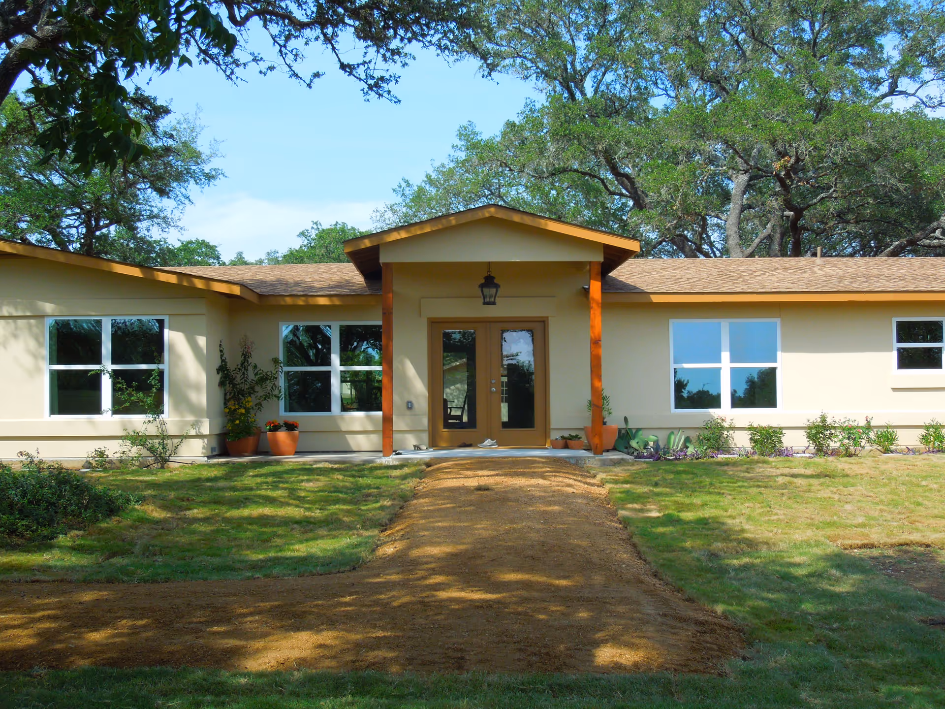 Front exterior view of a single-story building with a beige facade and brown roof. The entrance has double glass doors under a small covered porch supported by wooden posts. There are several windows on either side of the entrance and potted plants near the doorway. The building is surrounded by grass and trees.