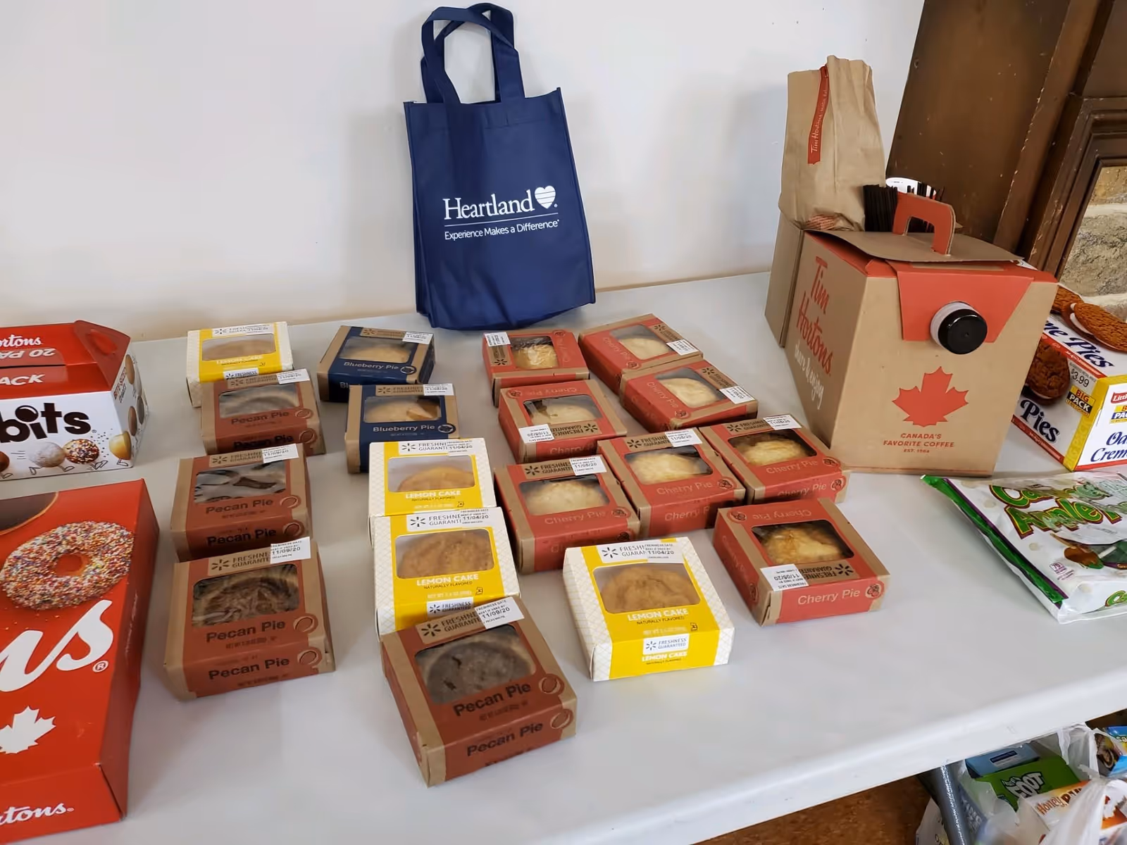 A table displaying various packaged baked goods including pecan pies, cherry pies, lemon cakes, blueberry pies, and a large box of Tim Hortons coffee. There is also a blue reusable bag with the Heartland logo and slogan, and some other snack boxes on the table.