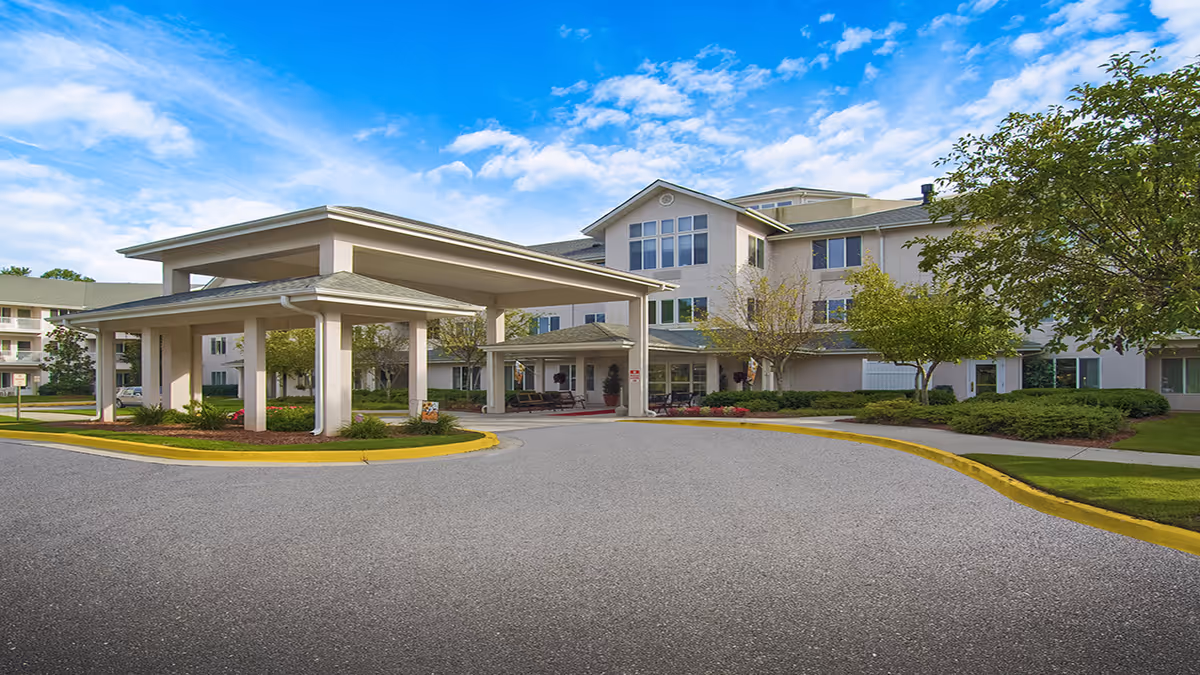 Front exterior view of Smoky Springs by Barclay House, showing a large covered entrance with pillars, a circular driveway, landscaped greenery, and a multi-story building under a partly cloudy blue sky.