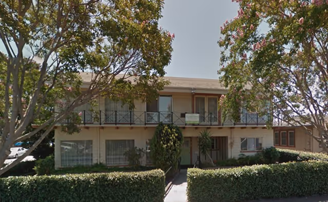 Two-story residential building with a balcony on the upper floor, surrounded by trees and hedges, under a clear sky.