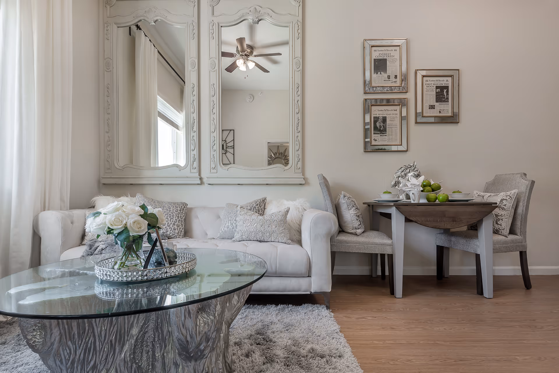 A cozy living and dining area featuring a white tufted sofa with decorative pillows, a glass coffee table with a silver tray holding white roses, and a small dining table set with green apples and a silver horse head sculpture. Two ornate mirrors hang above the sofa, and three framed newspaper-style prints are on the wall above the dining table. The room has light-colored walls, wood flooring, and a ceiling fan reflected in the mirrors.