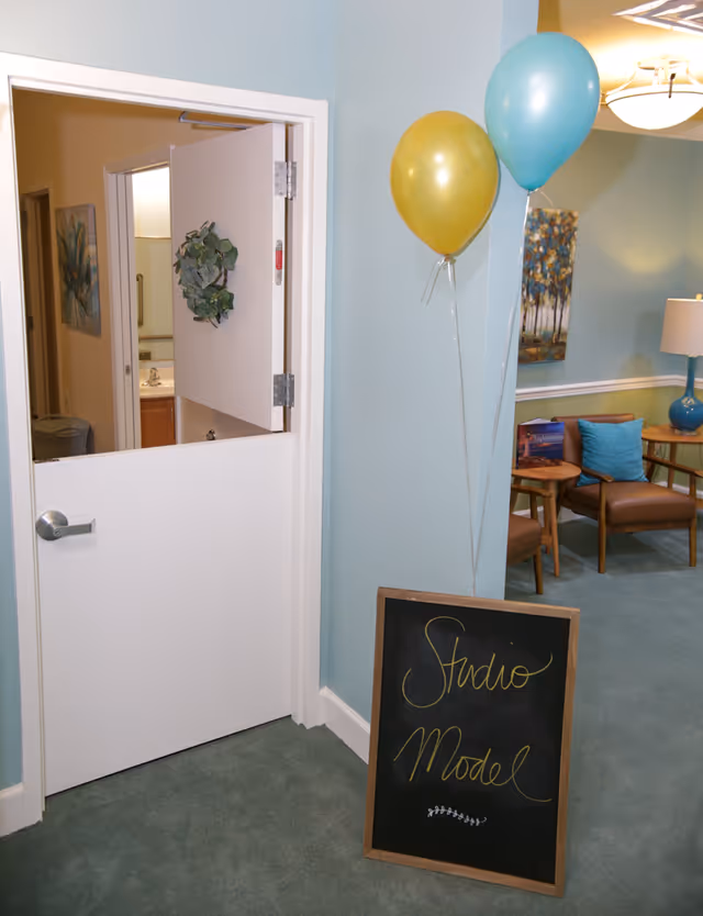 Interior view of a senior living facility hallway with a half-open white door decorated with a small green wreath. Next to the door, two balloons, one gold and one blue, are tied to the wall. A chalkboard sign on the floor reads 'Studio Model' in yellow cursive writing. In the background, there are brown chairs with blue cushions, a side table with magazines, a blue lamp, and a painting of trees on the wall.