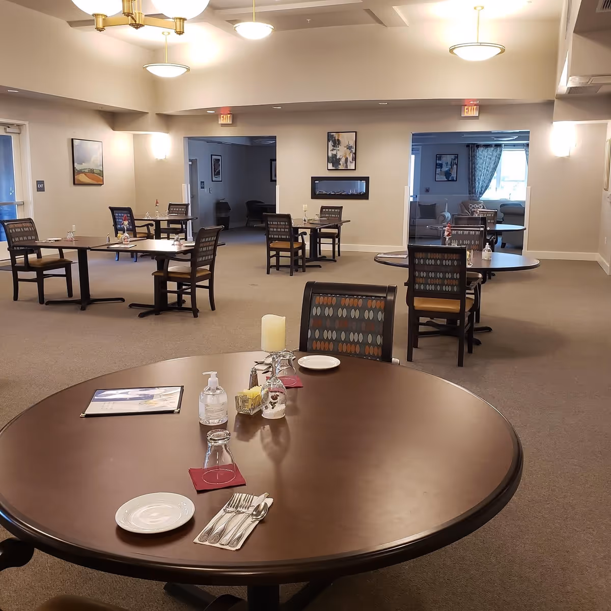 A spacious dining room with several round and square tables, each set with plates, utensils, glasses, and condiments. The room has beige walls, carpeted floors, and modern light fixtures hanging from the ceiling. There are paintings on the walls and an electric fireplace in the center wall. The room appears clean and well-lit with natural light coming from windows in an adjoining room.