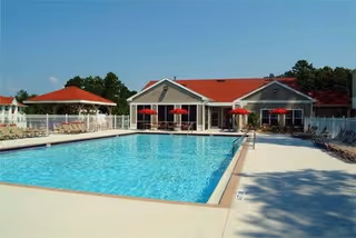 A large outdoor swimming pool with lounge chairs and a red‑roofed clubhouse and red umbrellas in the background.