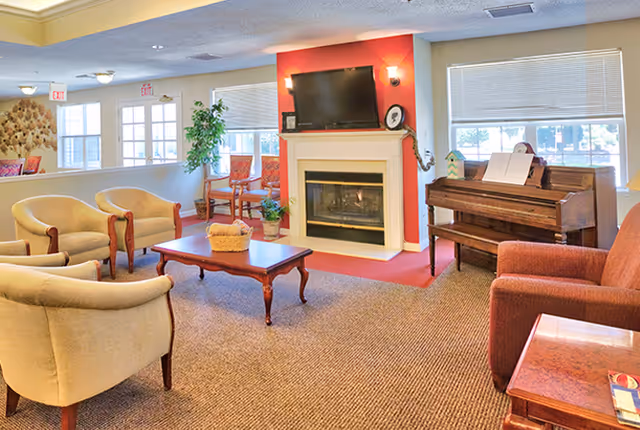 A cozy living room area in a senior living facility with beige armchairs, a wooden coffee table with a basket, a fireplace with a mounted TV above it, a piano with sheet music, and large windows letting in natural light.