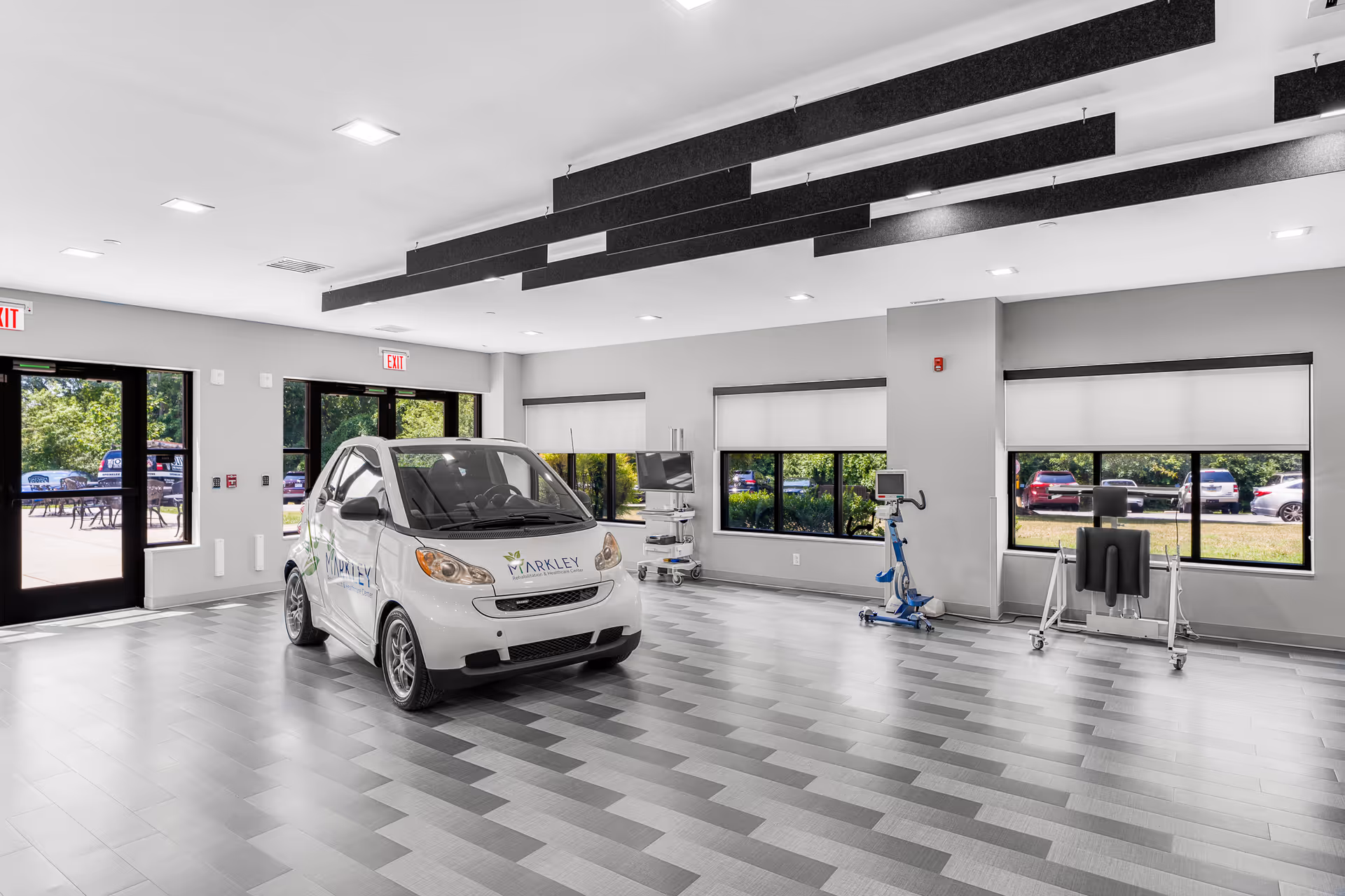 A spacious, modern indoor room with large windows showing a parking lot outside. Inside, there is a small white car with the Markley logo on the hood and doors. The room has a patterned floor, ceiling lights, and some medical or rehabilitation equipment along the walls.