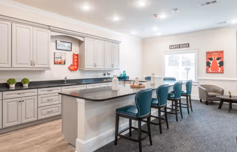 A bright and modern kitchen area with white cabinetry, a long black countertop island with four teal upholstered bar stools, and decorative wall art including a 'Happy Hour' sign above a window. There is also a small seating area with a chair and a side table near the window.