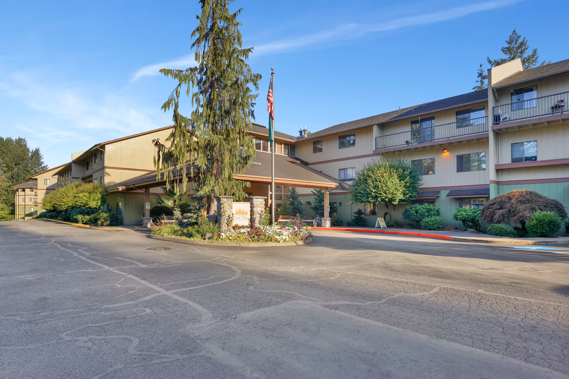 Exterior view of The Firs senior living facility showing a multi-story building with balconies, surrounded by trees and shrubs, with a flagpole and a sign at the entrance under a clear blue sky.