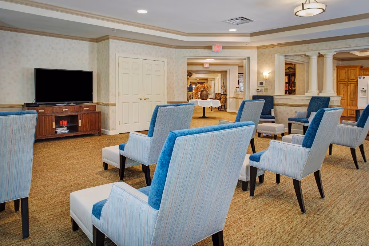 A common area in a senior living facility with multiple blue and beige upholstered chairs arranged facing a large flat-screen TV on a wooden cabinet. The room has beige patterned wallpaper, carpeted floor, and recessed lighting. There is a table with a floral arrangement in the background and an open doorway leading to other parts of the facility.