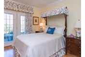 Sunlit bedroom with a canopy-topped bed, wooden nightstands with lamps, and French doors leading outside.