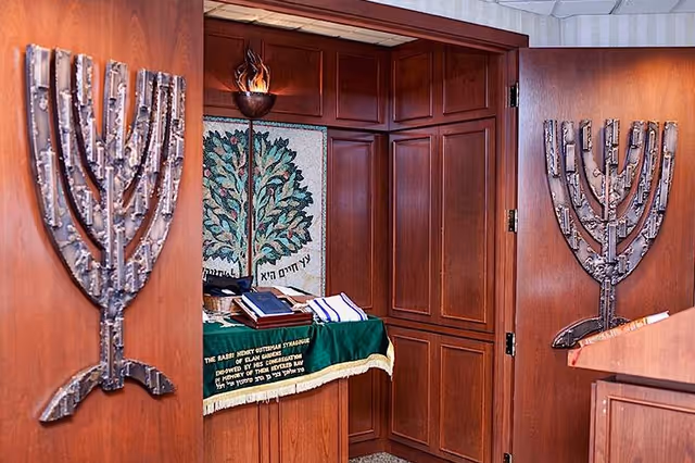 Interior view of a small synagogue or prayer room with wooden walls and doors. Two large metal menorahs are mounted on the wooden doors. Inside the room, there is a table covered with a green cloth, holding religious books and a tall torch-like candle holder. A tapestry with a tree design and Hebrew text hangs on the back wall.