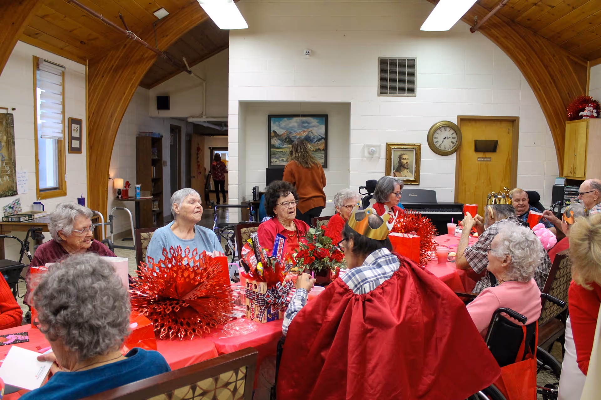 A group of elderly people gathered around a long table covered with a red tablecloth, decorated with red festive decorations and gift bags. Some individuals are wearing paper crowns and engaging in conversation in a cozy room with wooden arched ceilings and white walls. A clock, paintings, and a piano are visible in the background.